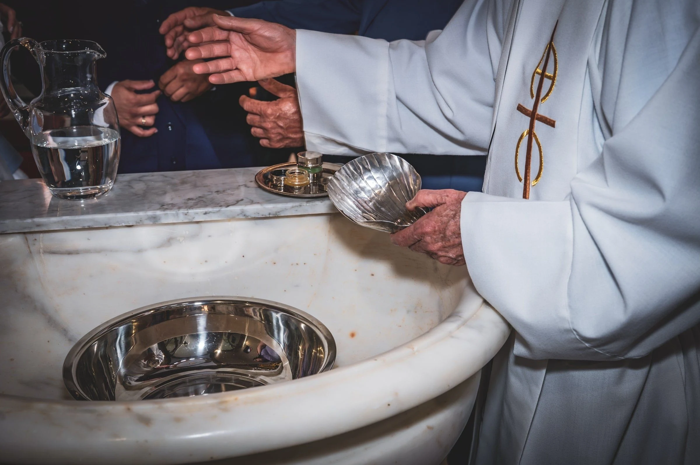 Manos del sacerdote con una concha plateada junto a la jarra de agua y la pila bautismal.