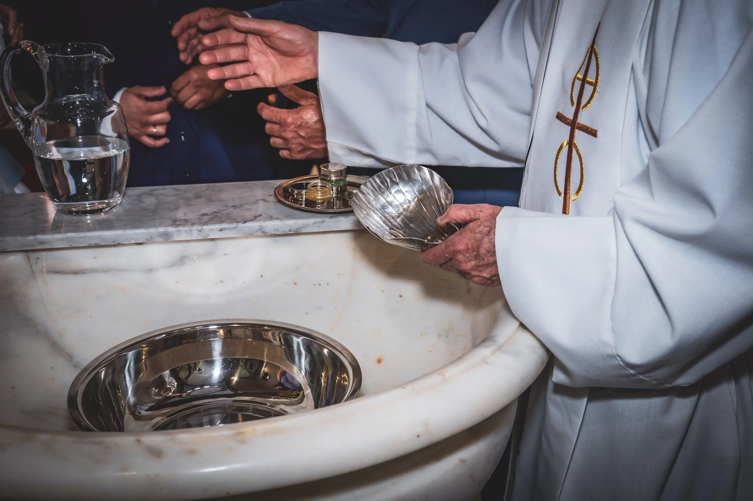 Manos del sacerdote con una concha plateada junto a la jarra de agua y la pila bautismal.