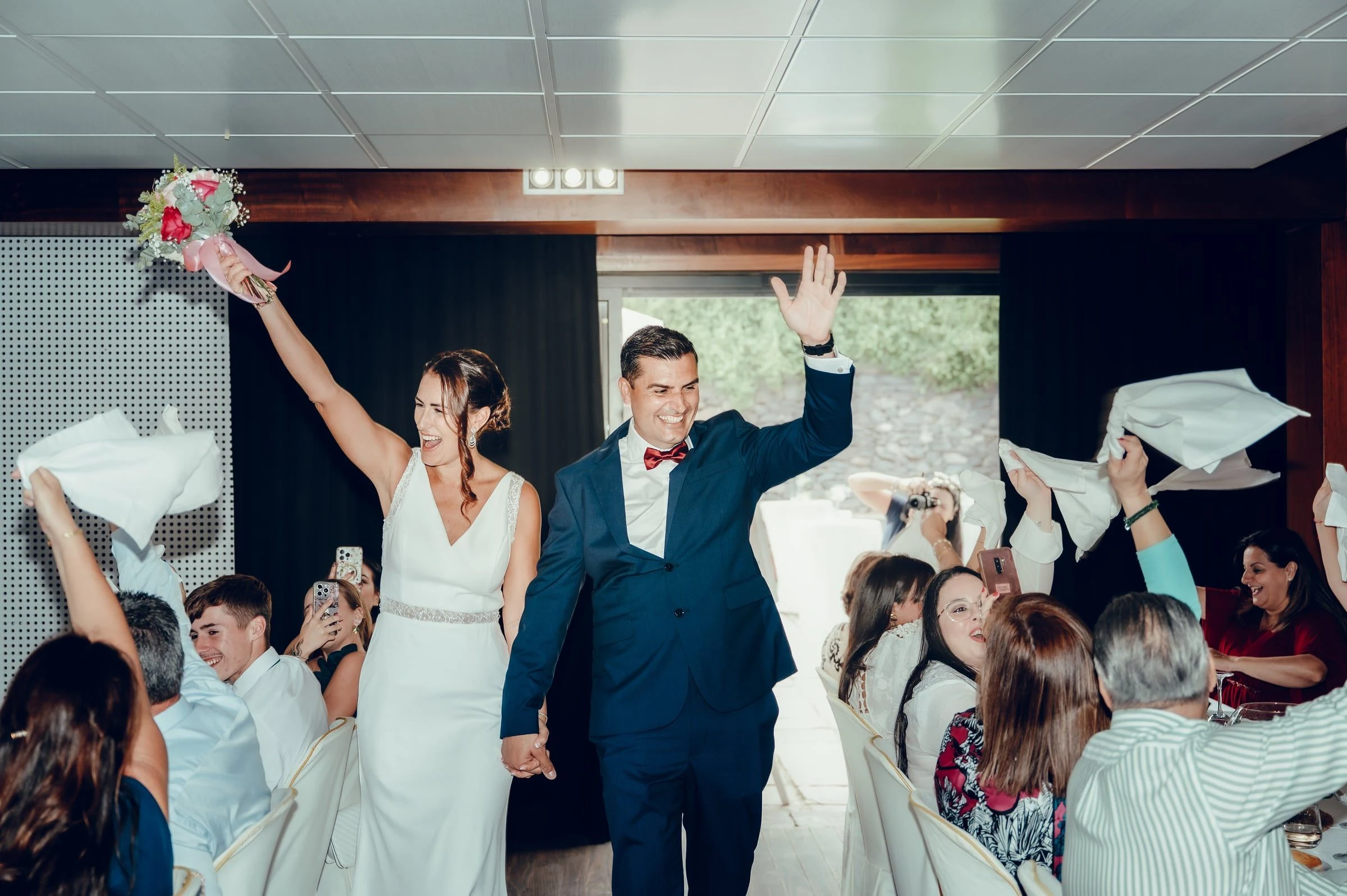 Novios levantando los brazos entre invitados que agitan servilletas en el banquete.