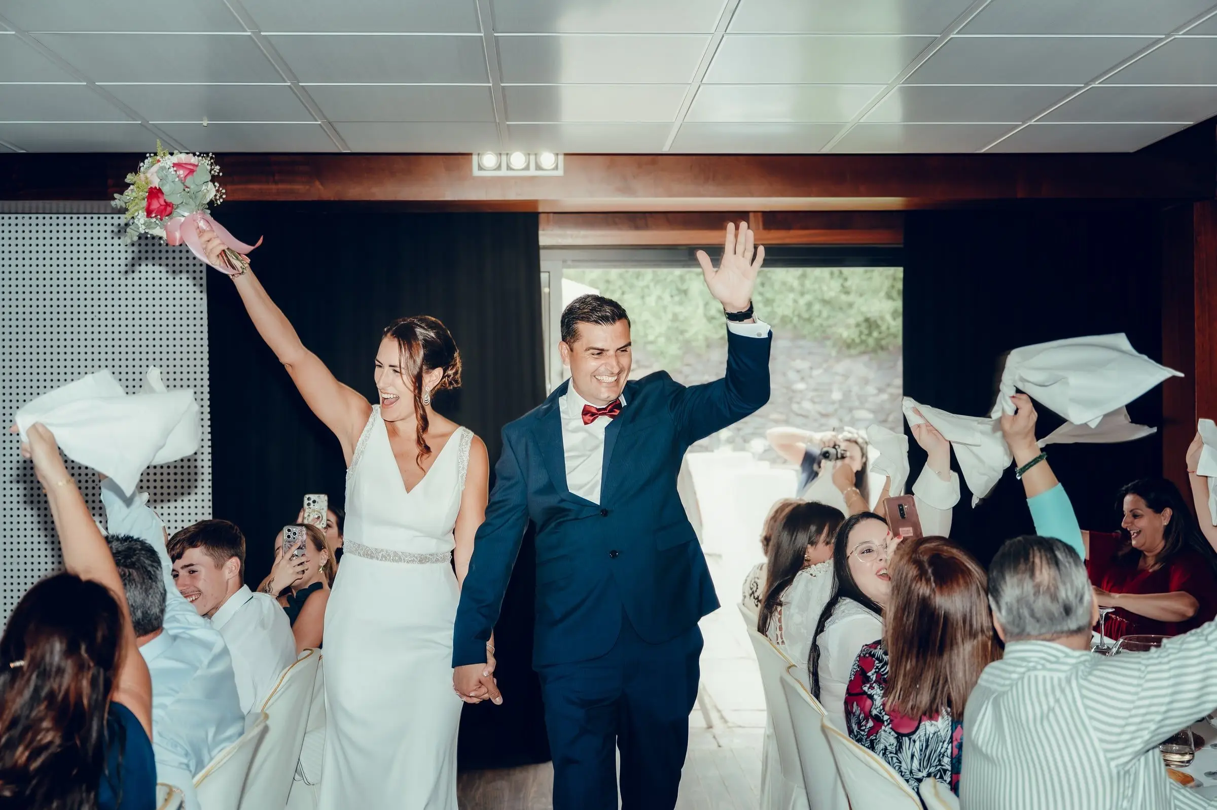 Novios levantando los brazos entre invitados que agitan servilletas en el banquete.