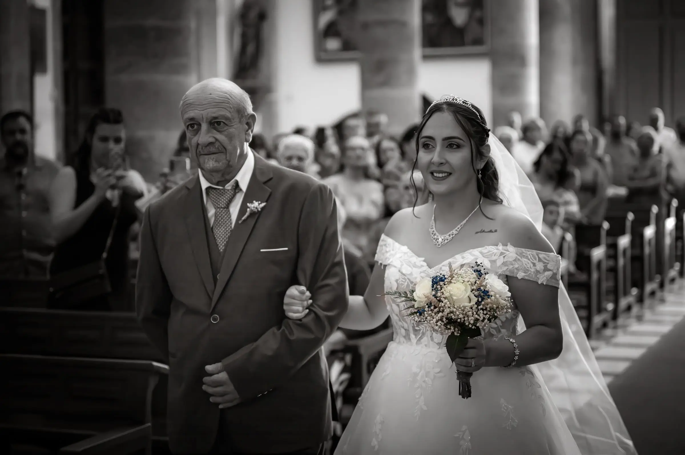 Novia entrando del brazo de un acompañante entre los invitados, en blanco y negro.