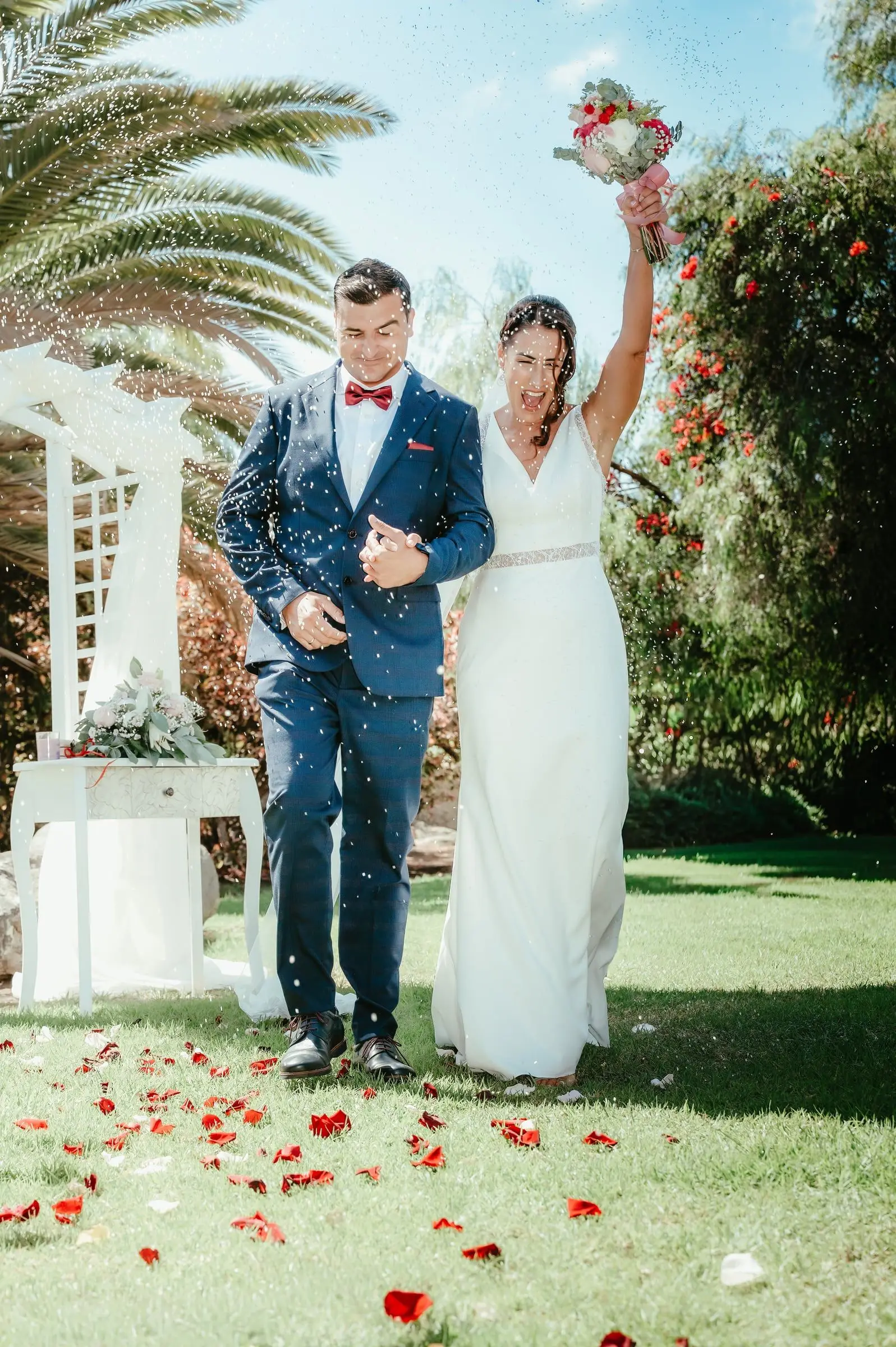 Pareja de novios avanzando de la mano entre pétalos y palmeras.