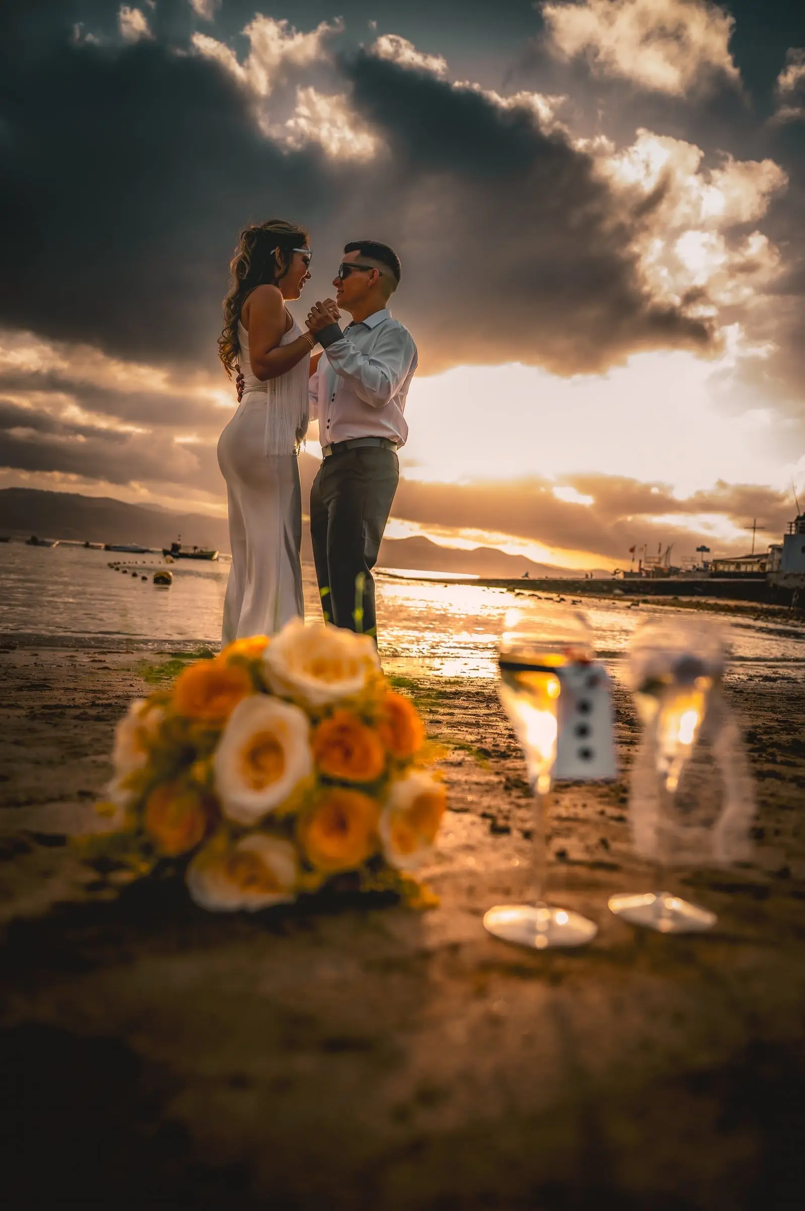 Pareja de novios abrazada junto al mar al atardecer, con un ramo amarillo en primer plano.