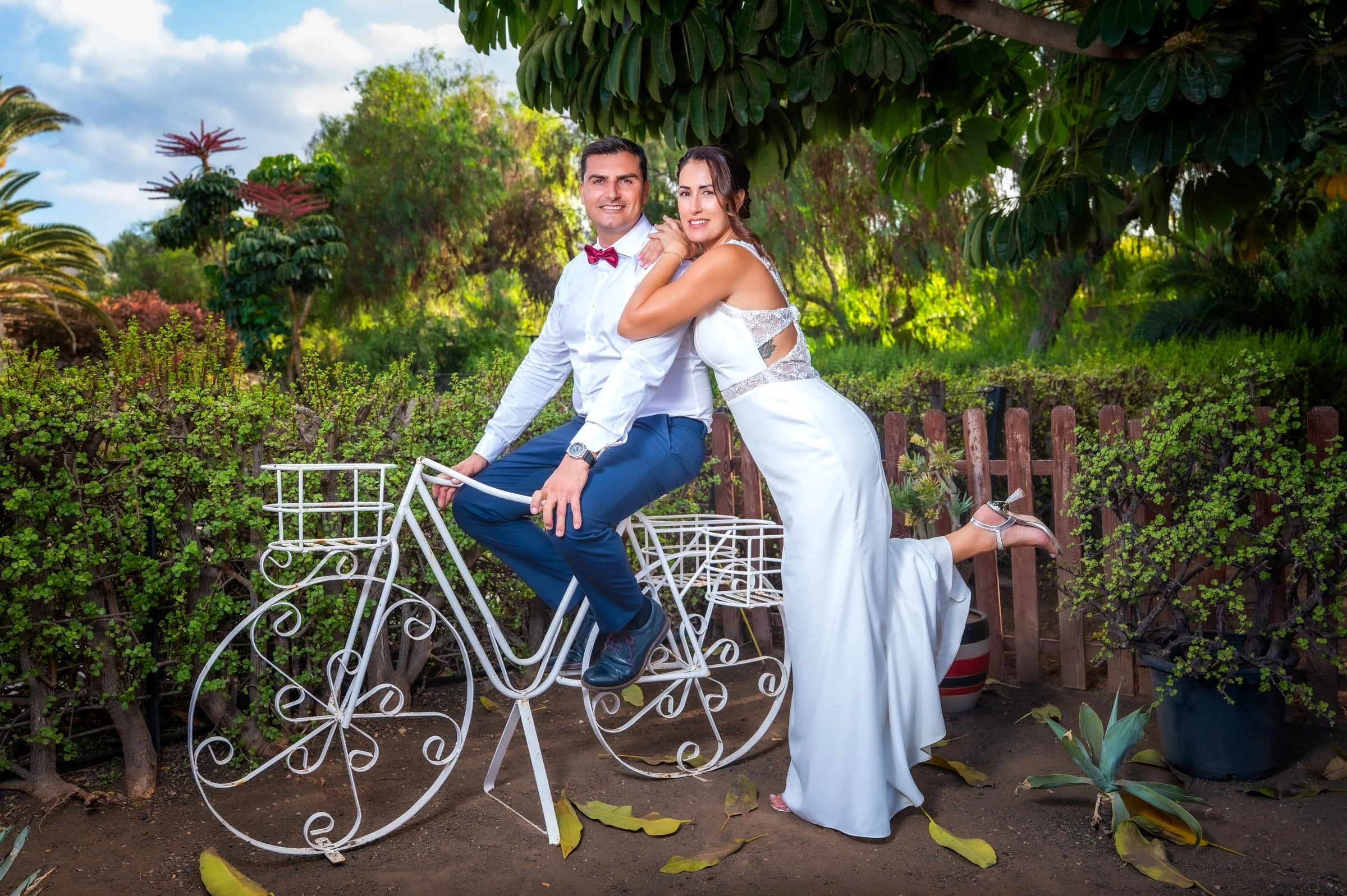 Pareja de novios junto a una bicicleta decorativa blanca rodeada de vegetación.