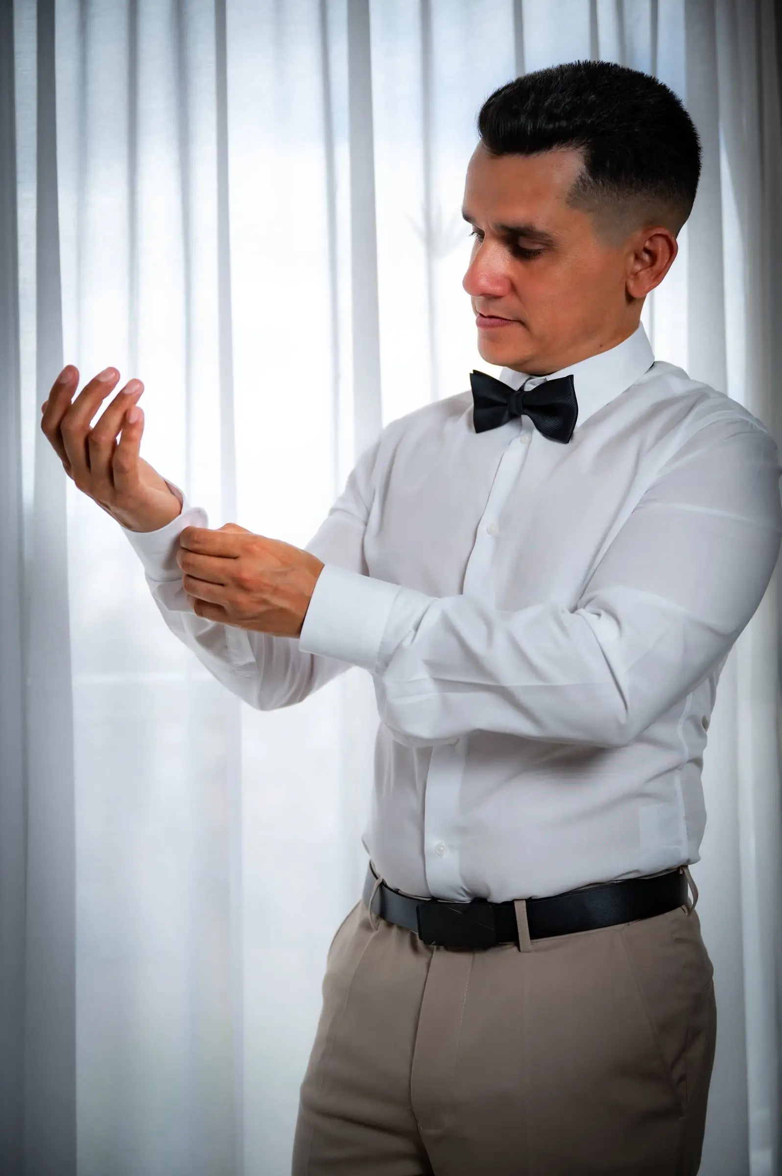 Novio ajustándose el puño de la camisa junto a una ventana.