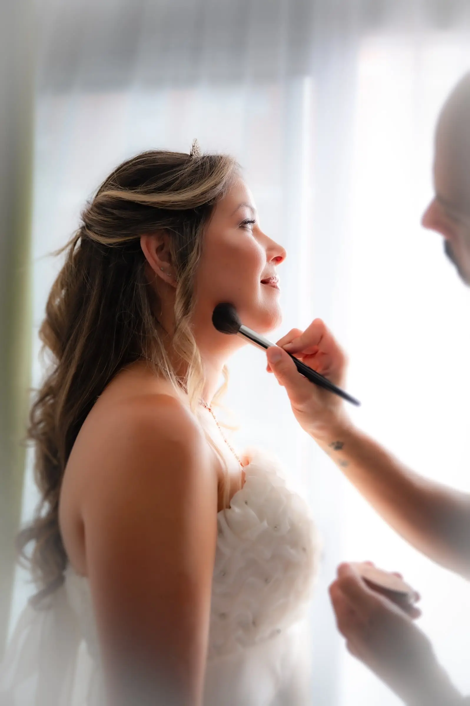 Novia de perfil mientras le retocan el maquillaje junto a la ventana.