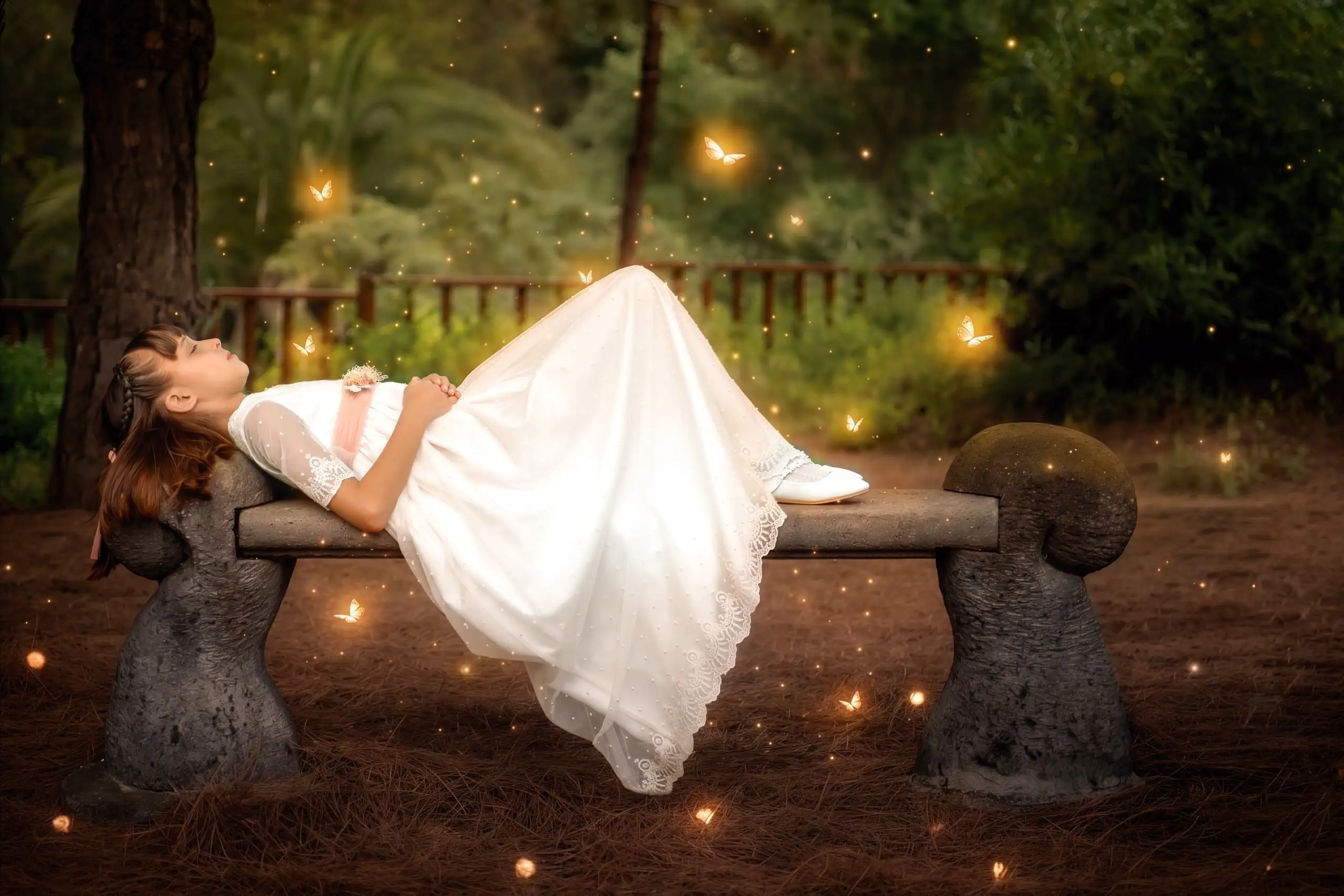 Niña con vestido de comunión recostada sobre un banco de piedra en un jardín.