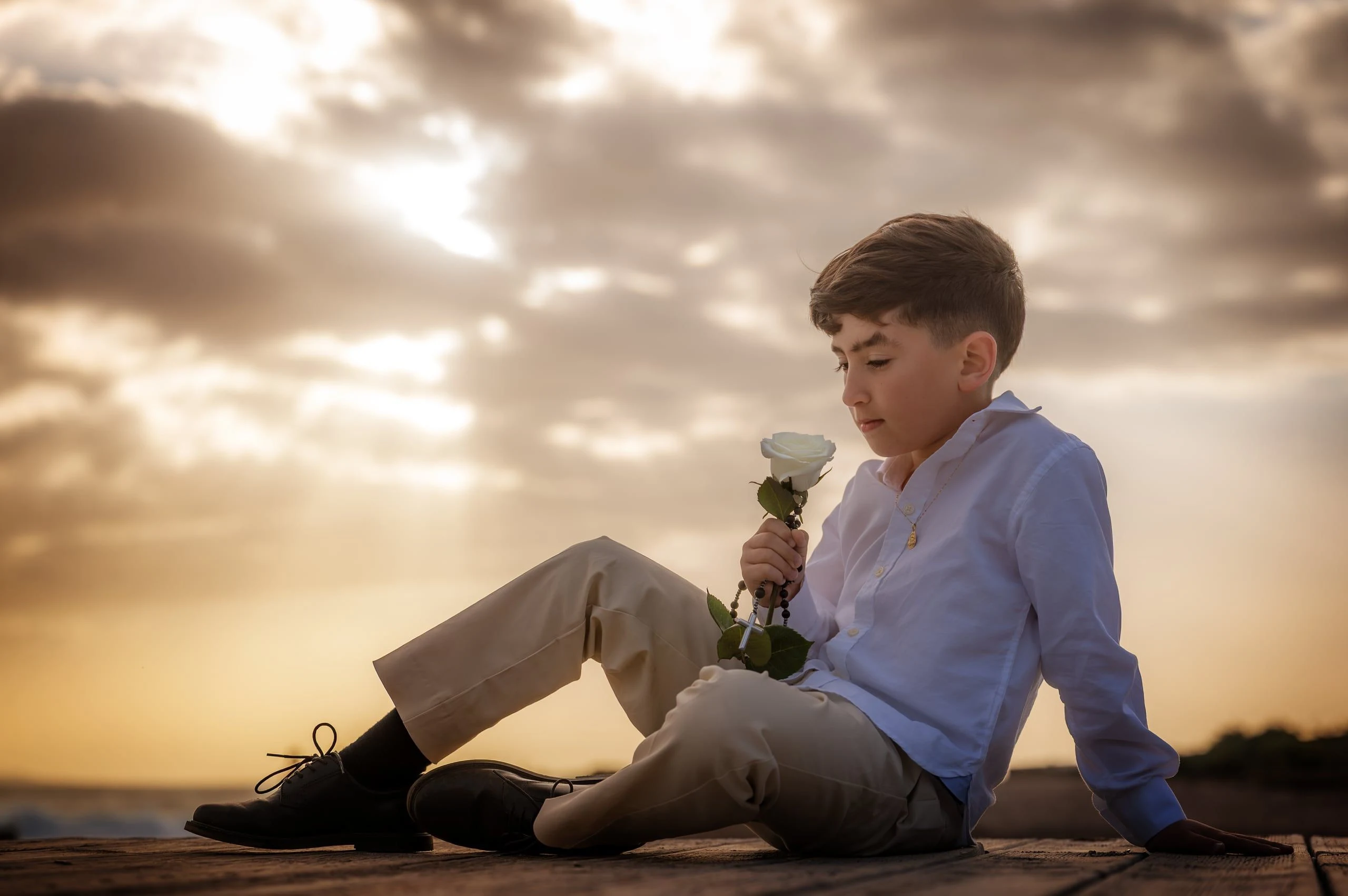 Niño de comunión sentado en la arena con una rosa al atardecer.