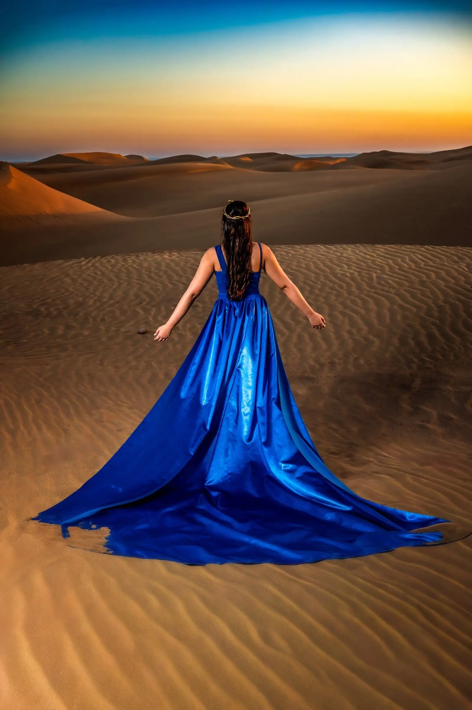 Quinceañera de espaldas con la cola de su vestido azul extendida sobre las dunas al atardecer.