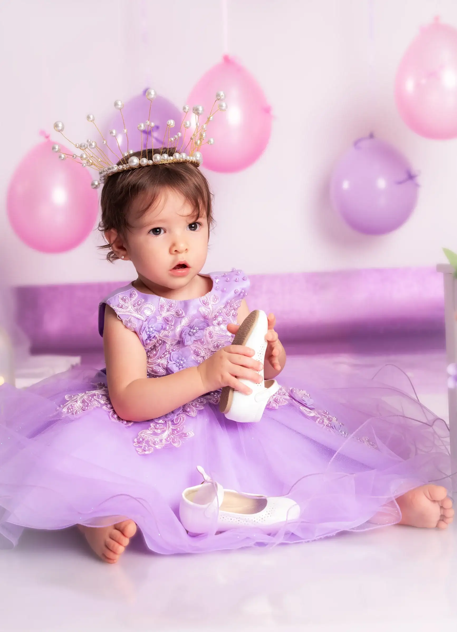 Niña pequeña con vestido lila sosteniendo un zapato entre las manos en el estudio.