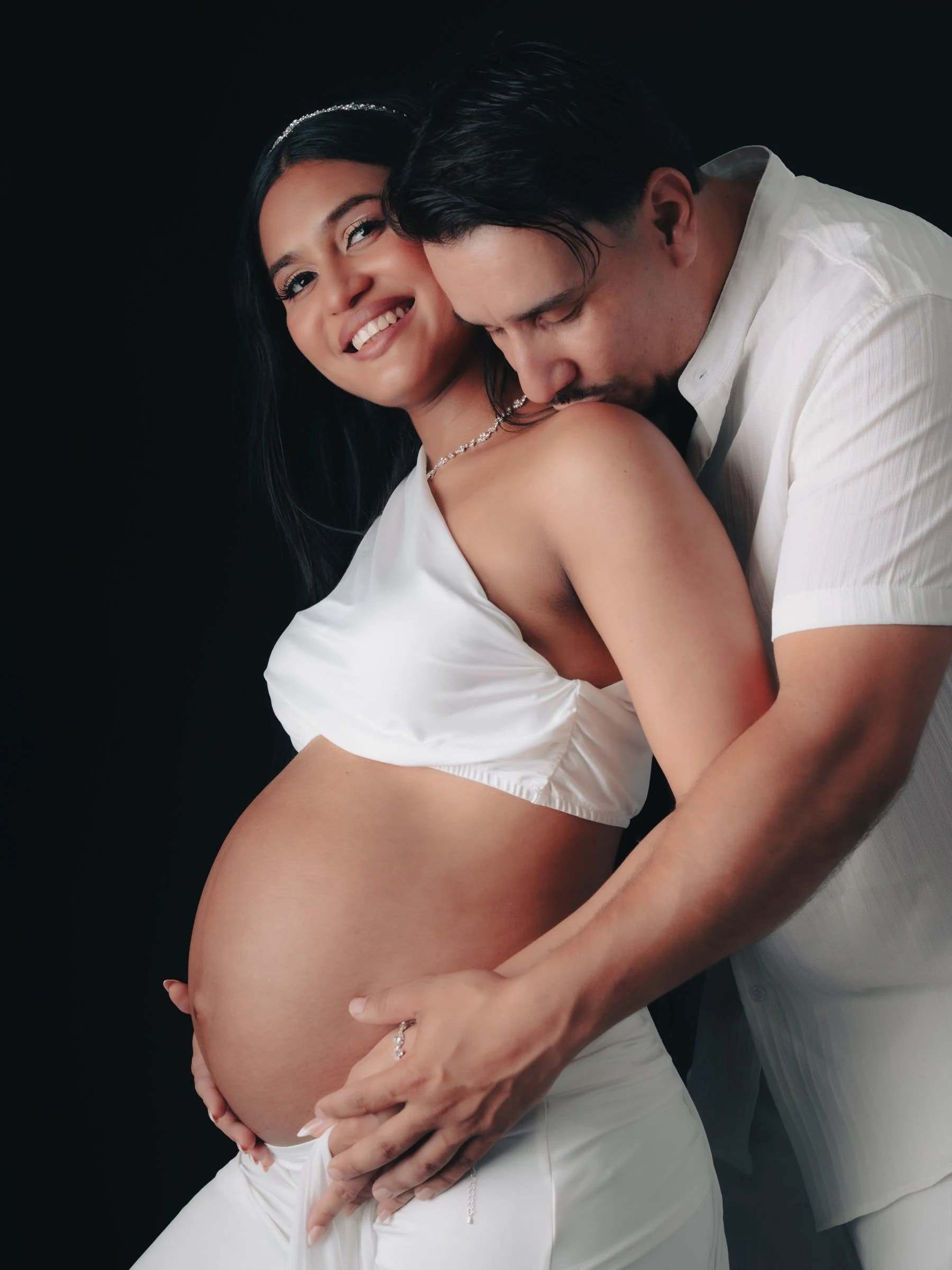Pareja abrazada junto a la barriga durante una sesión de embarazo con telas blancas.