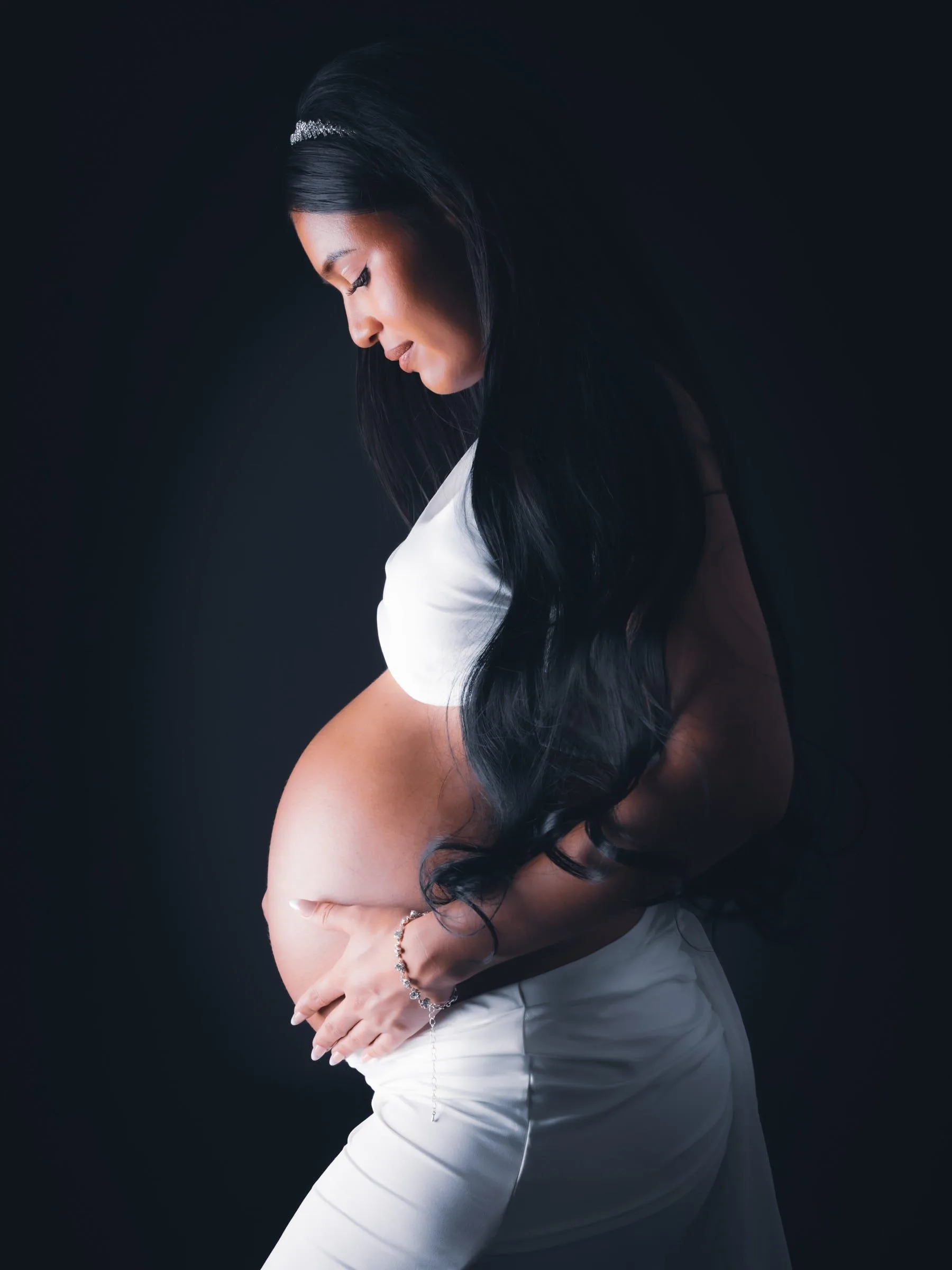 Mujer embarazada de perfil con las manos sobre la barriga y luz lateral suave.