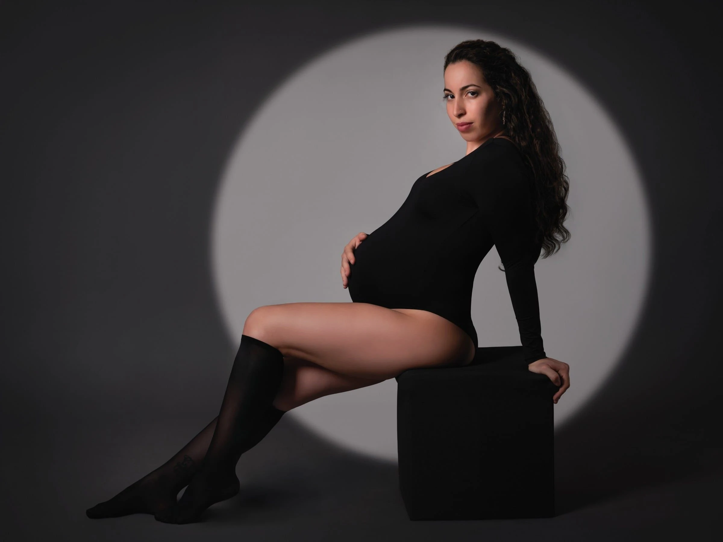 Mujer embarazada sentada de perfil con vestido negro ante un círculo de luz.