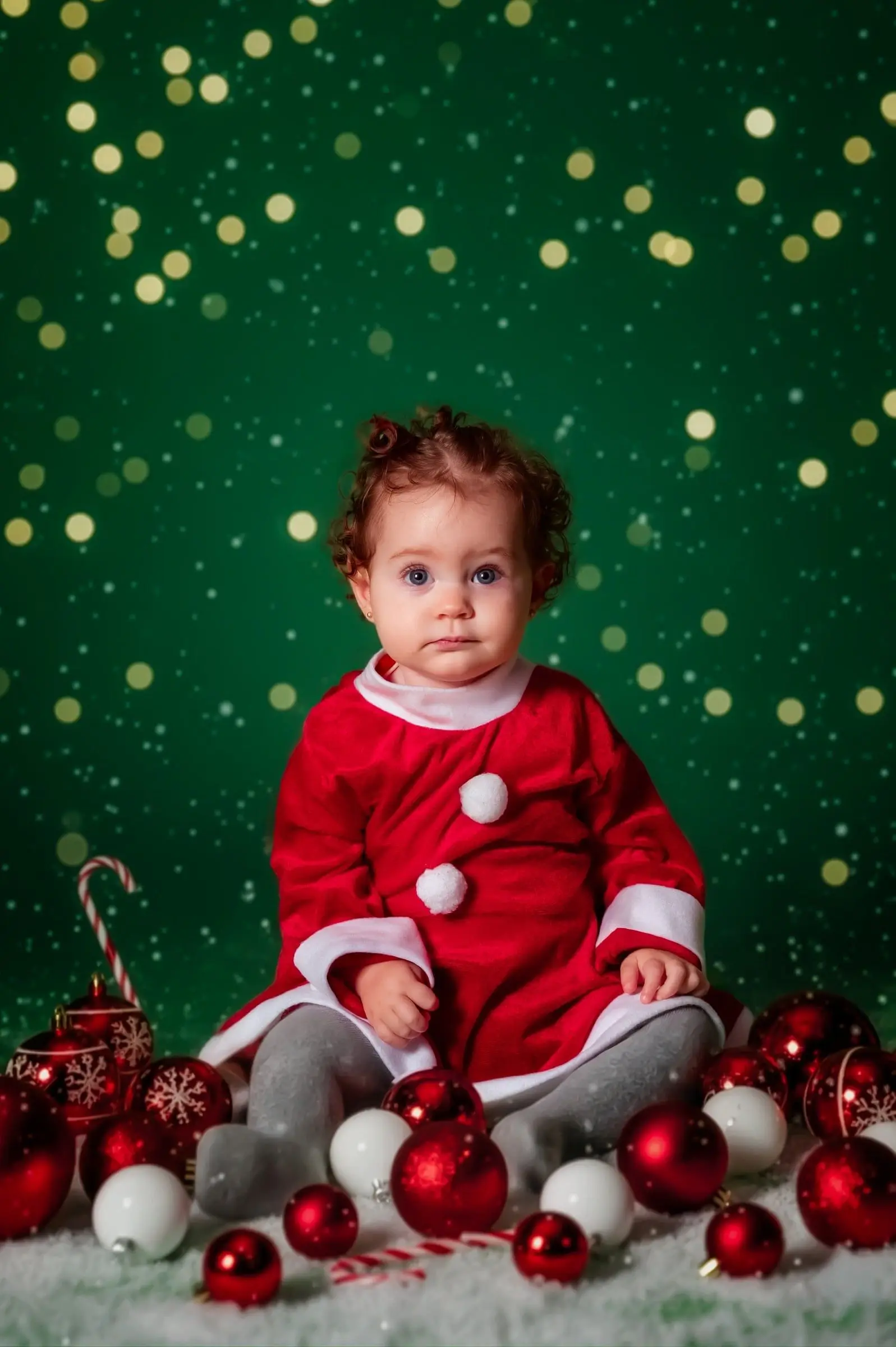 Bebé con ropa roja sentado ante un fondo verde con luces navideñas.