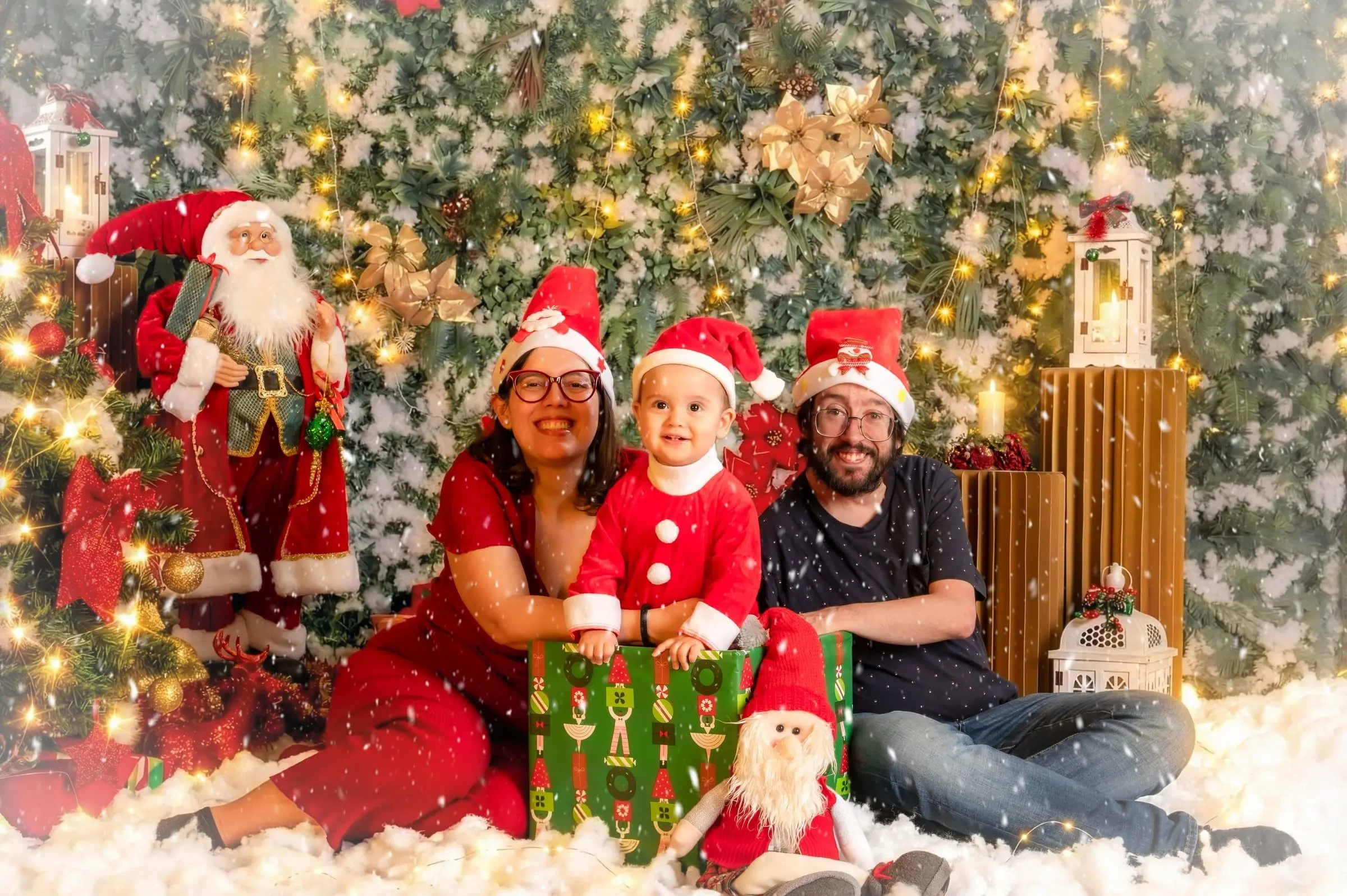 Dos adultos y un niño con gorro de Navidad sentados junto a un regalo verde.