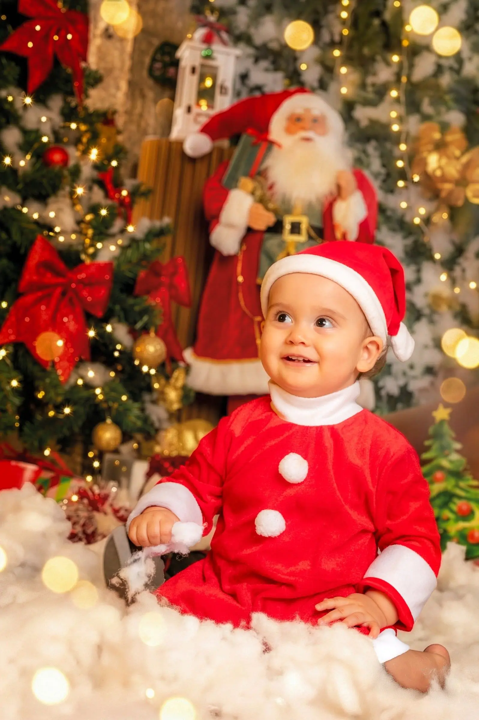 Bebé sonriente con gorro rojo sentado junto al árbol y un muñeco de Papá Noel.