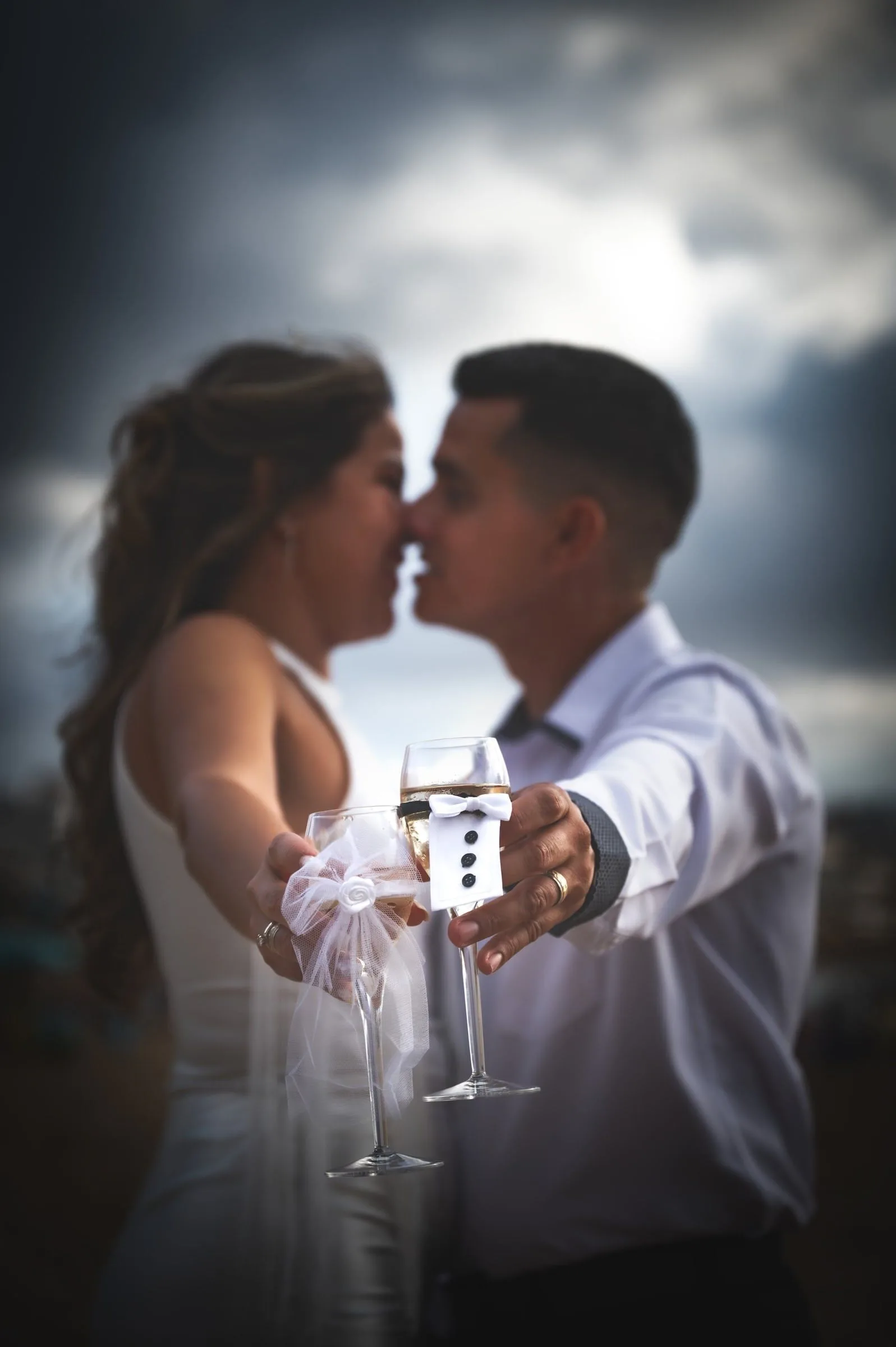 Pareja acercándose para un beso a contraluz, con dos copas de brindis en primer plano.