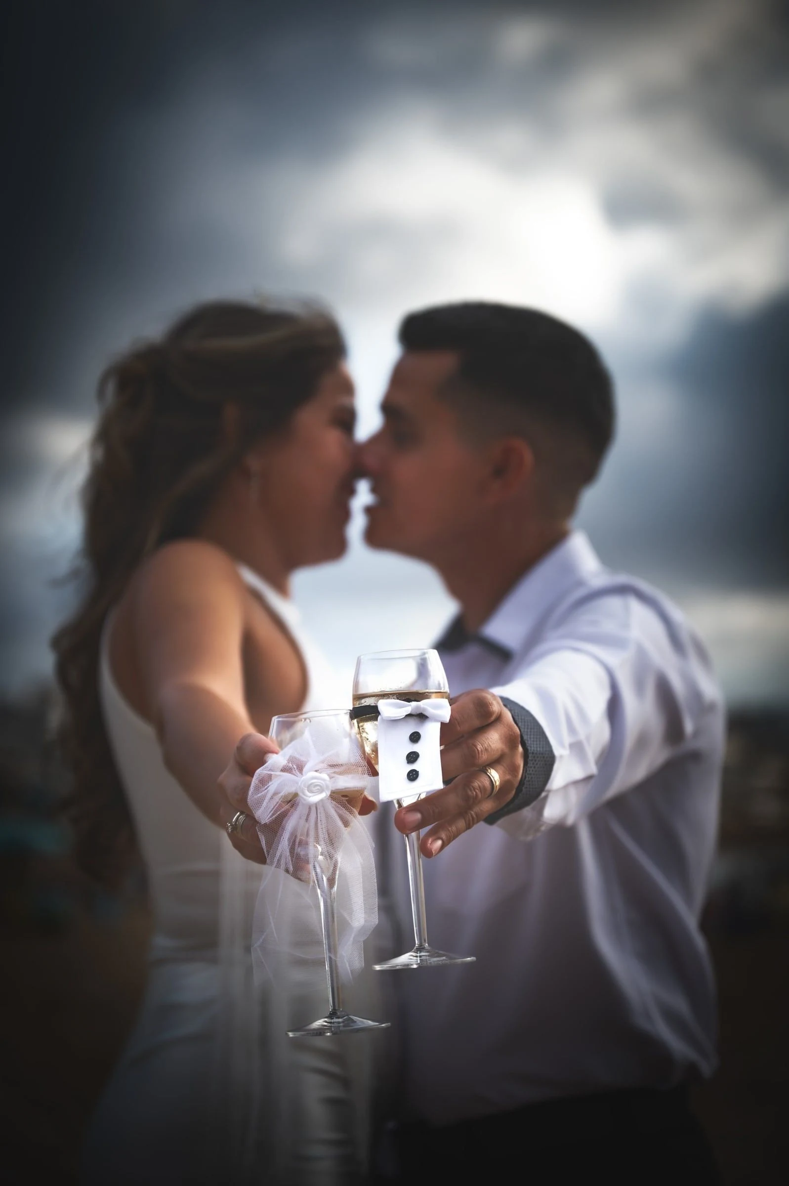 Pareja acercándose para un beso a contraluz, con dos copas de brindis en primer plano.