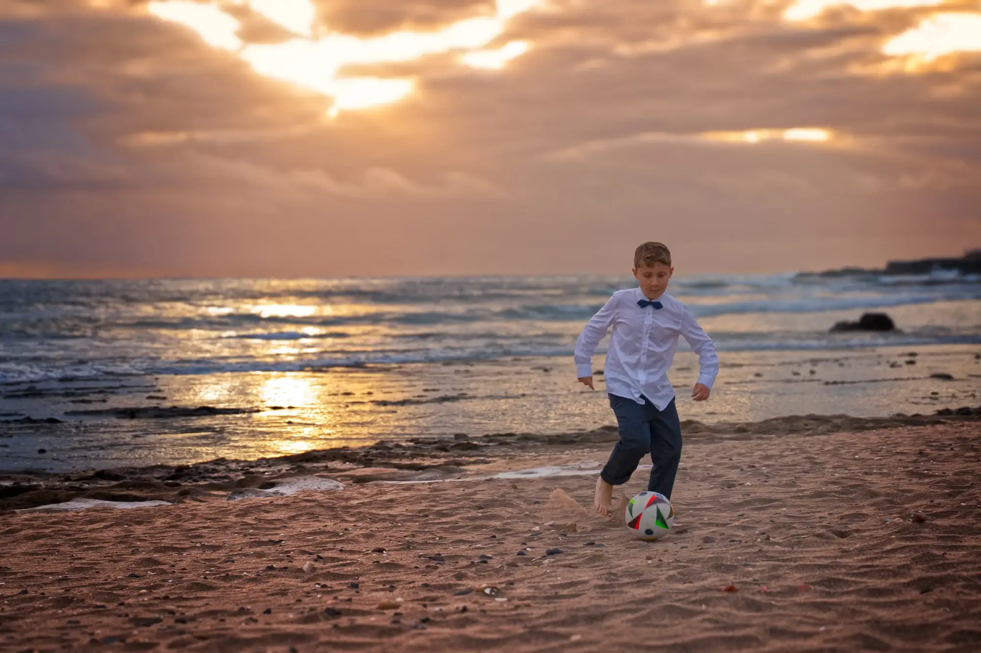 Niño jugando con un balón en la orilla al atardecer.