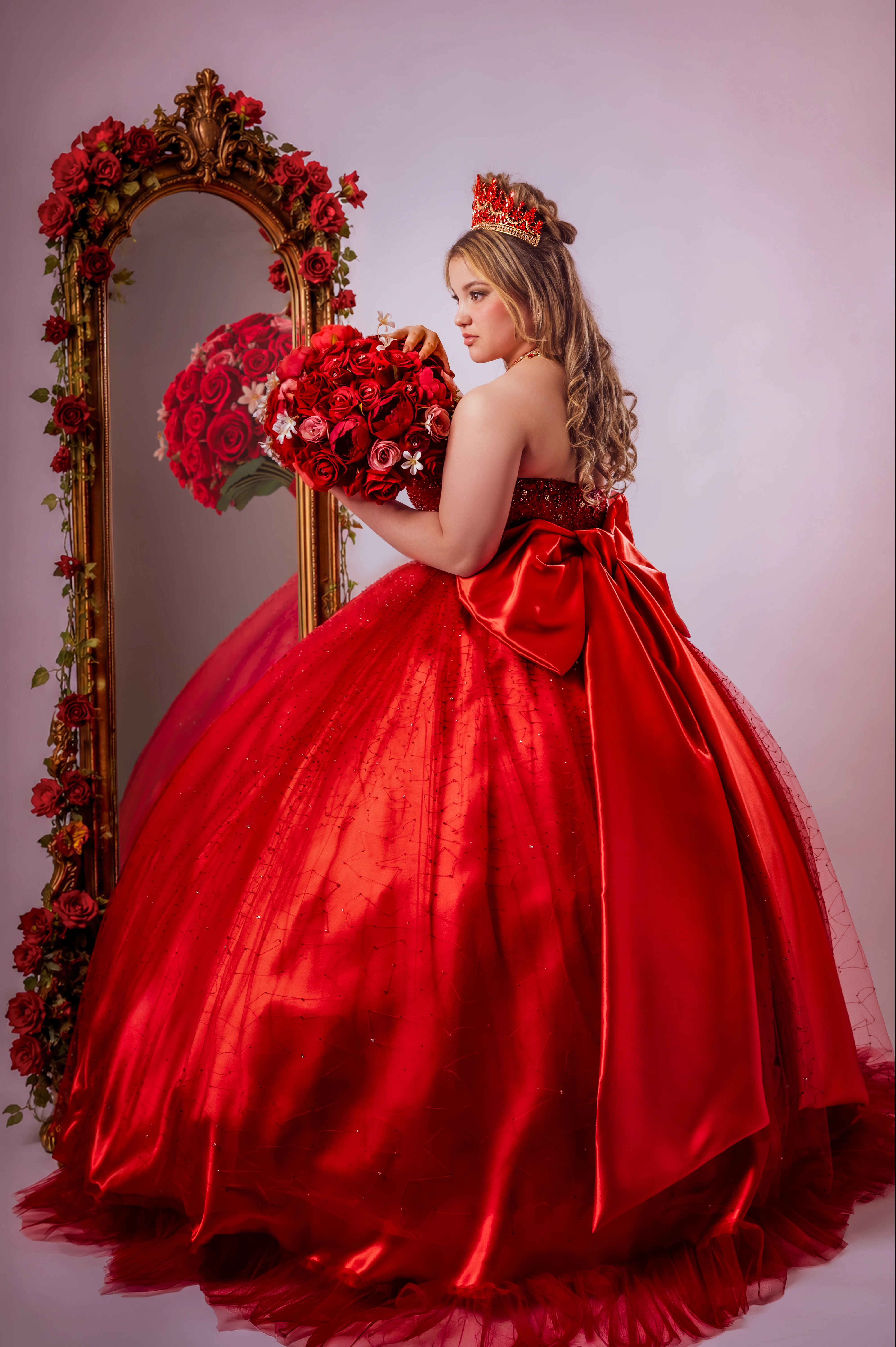 Quinceañera con vestido rojo y ramo de rosas frente a un espejo decorado.
