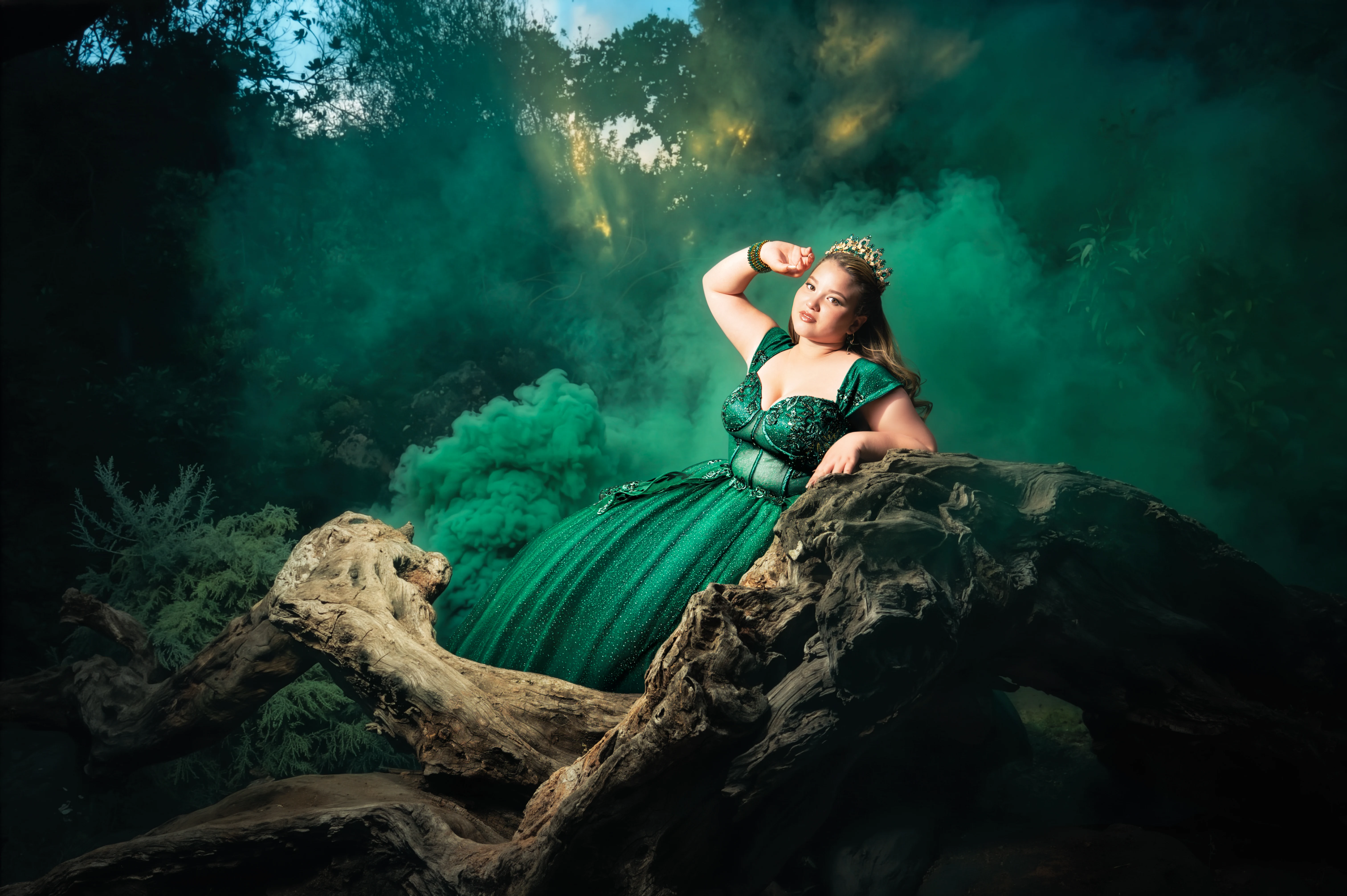 Quinceañera con vestido verde sobre un tronco entre humo y naturaleza.