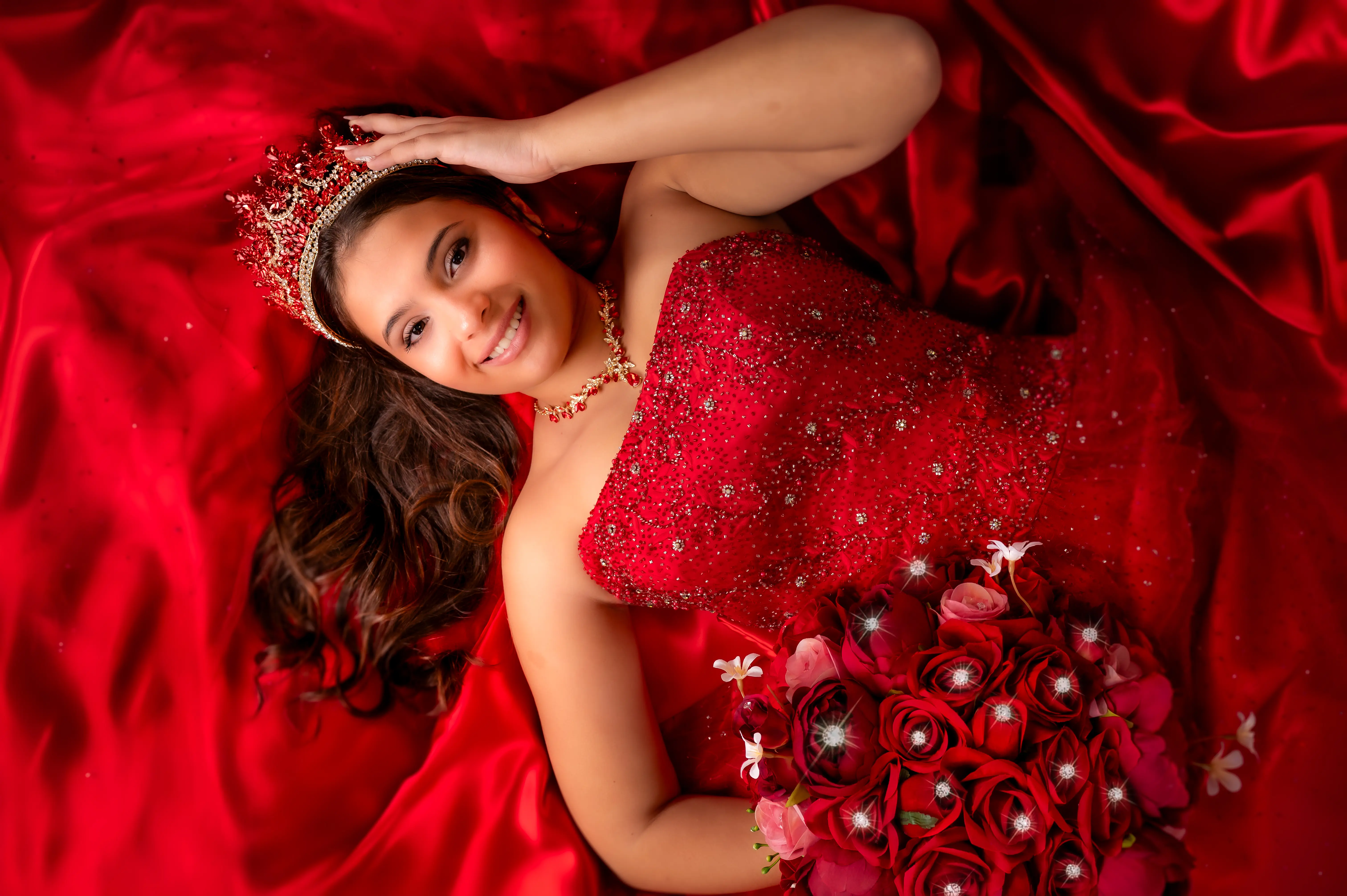 Quinceañera con vestido rojo recostada sobre tela roja y un ramo de flores.