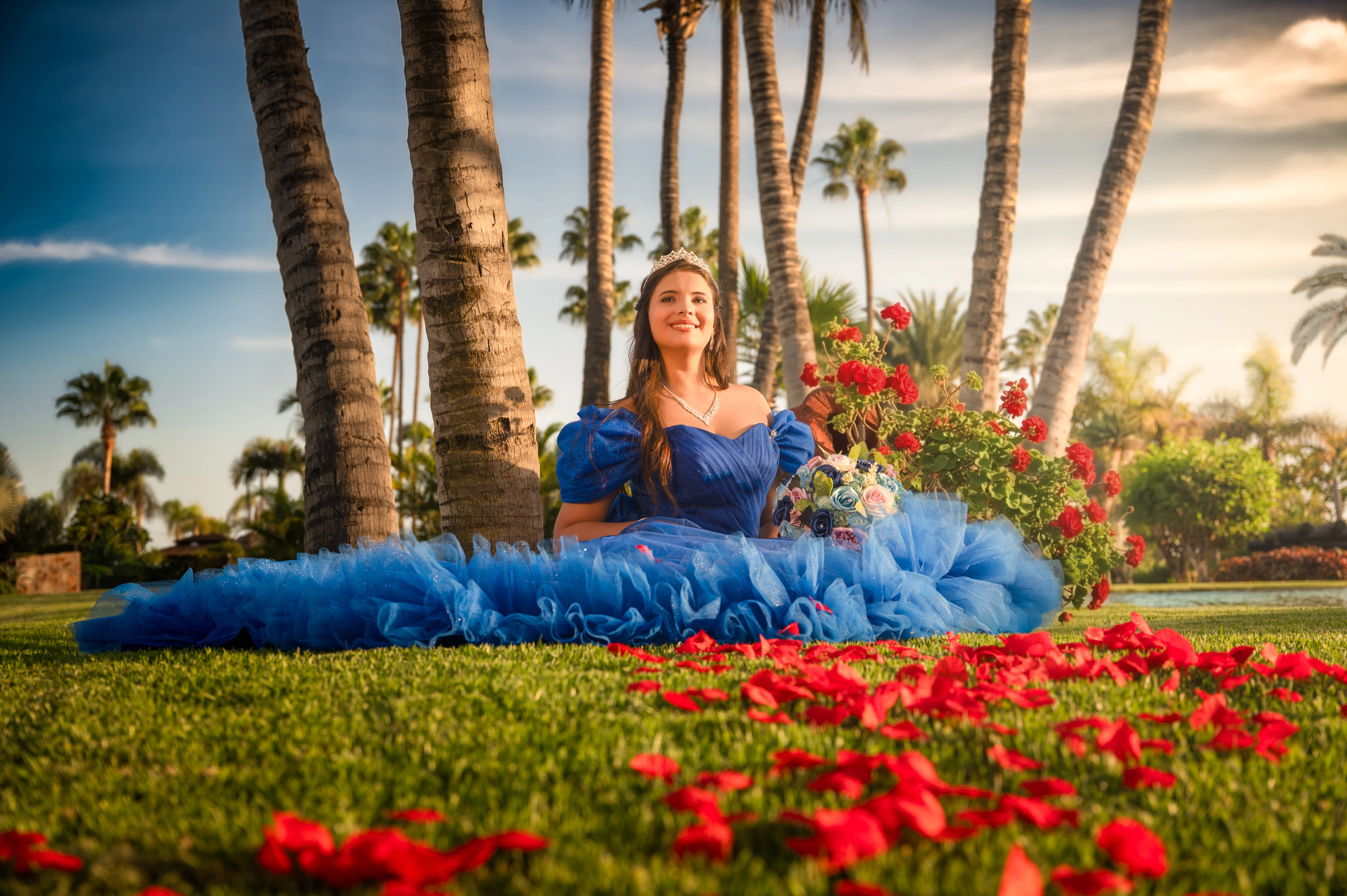 Quinceañera con vestido azul en un jardín de palmeras cubierto de flores rojas.