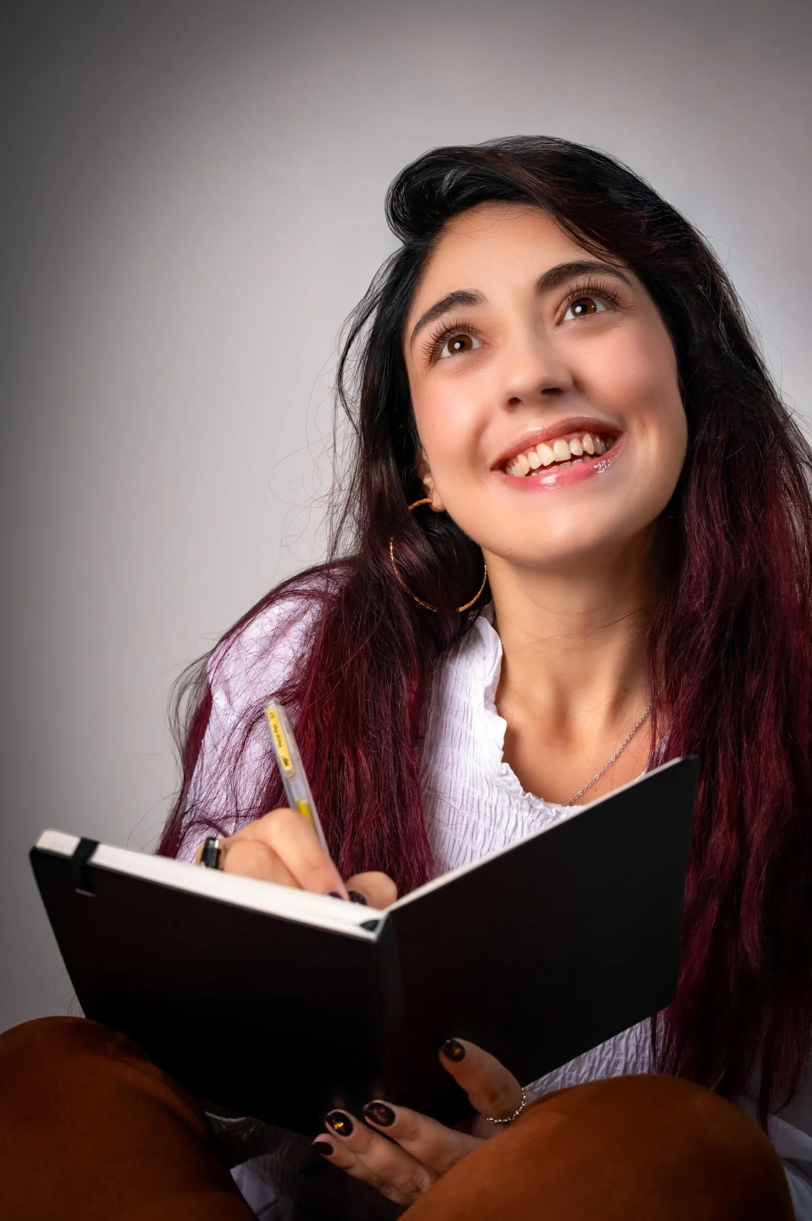 Mujer sonriente mirando hacia arriba, con un cuaderno abierto y un lápiz.