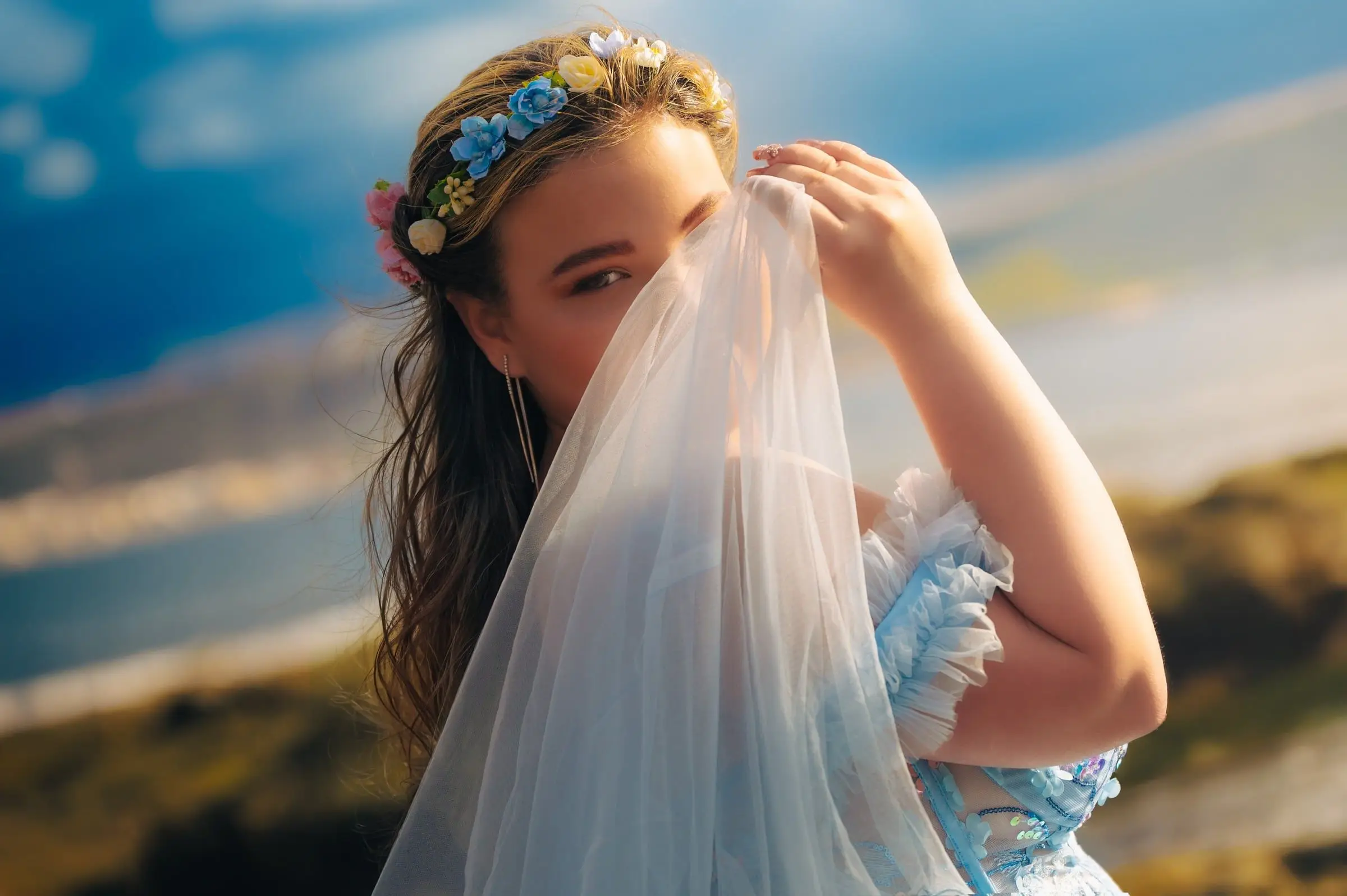 Retrato de quinceañera con una tela azul junto al rostro y el mar al fondo.
