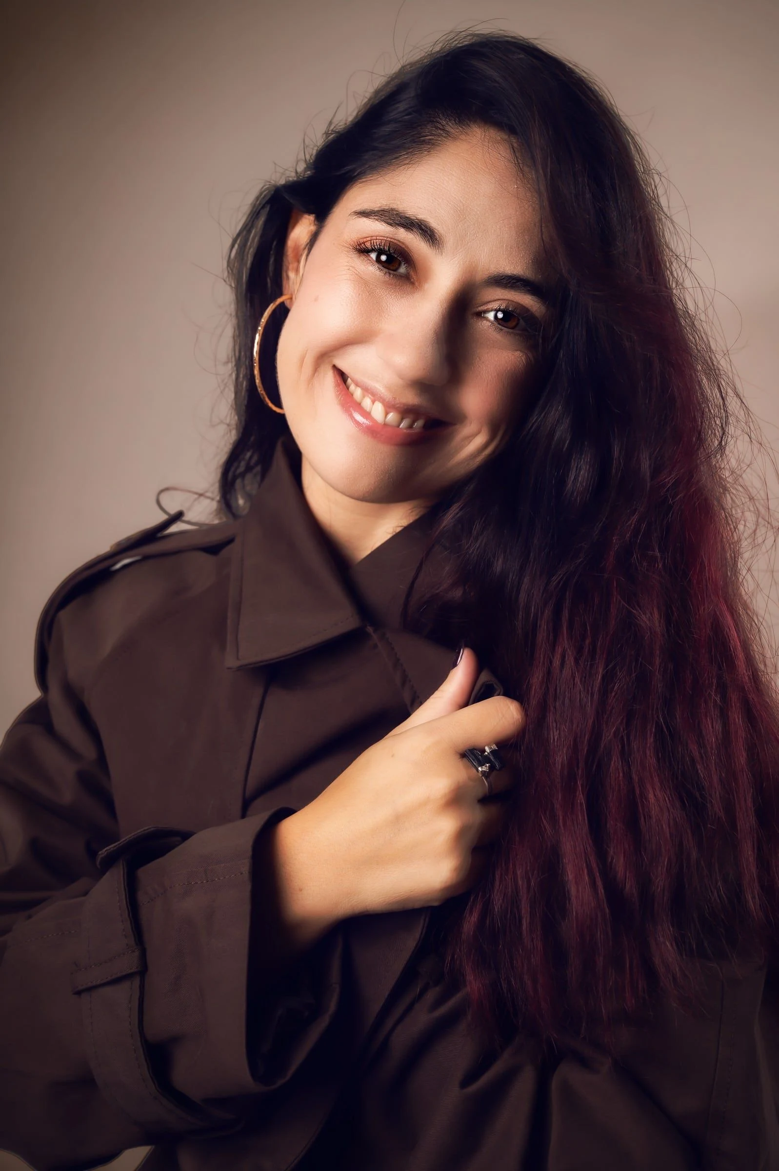 Mujer sonriente con camisa oscura y una mano junto al cabello, sobre fondo cálido.