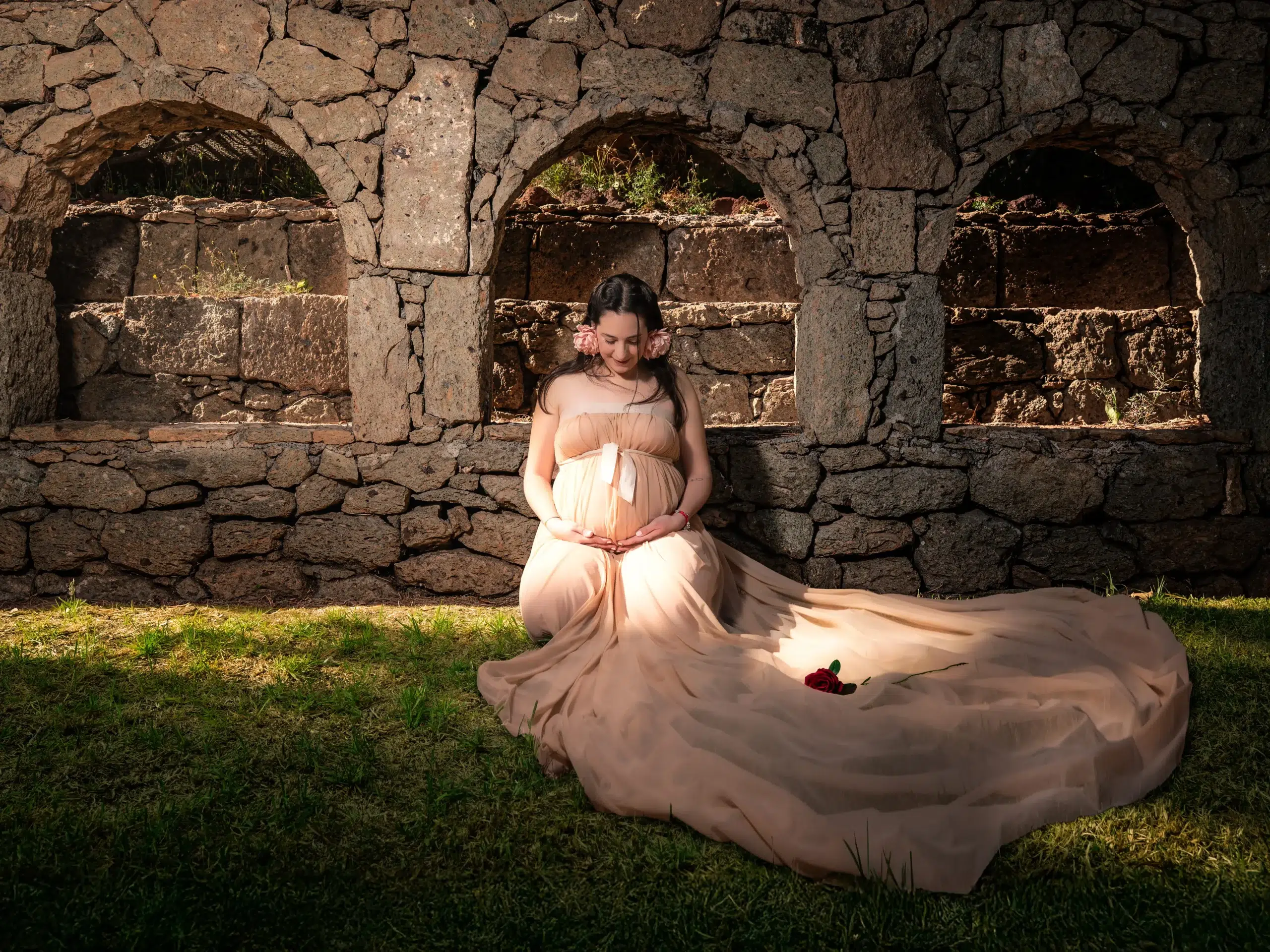 Mujer embarazada con vestido largo posando sentada ante una construcción de piedra durante una sesión fotográfica en Gran Canaria.