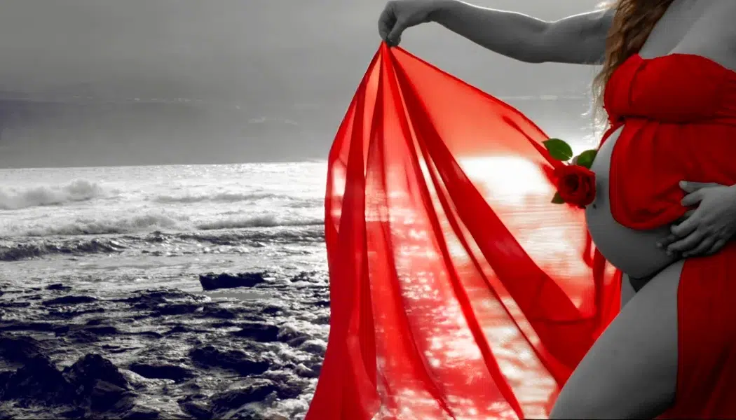 Fotografía artística de una mujer embarazada con vestido rojo y una rosa posando junto al mar en Gran Canaria.
