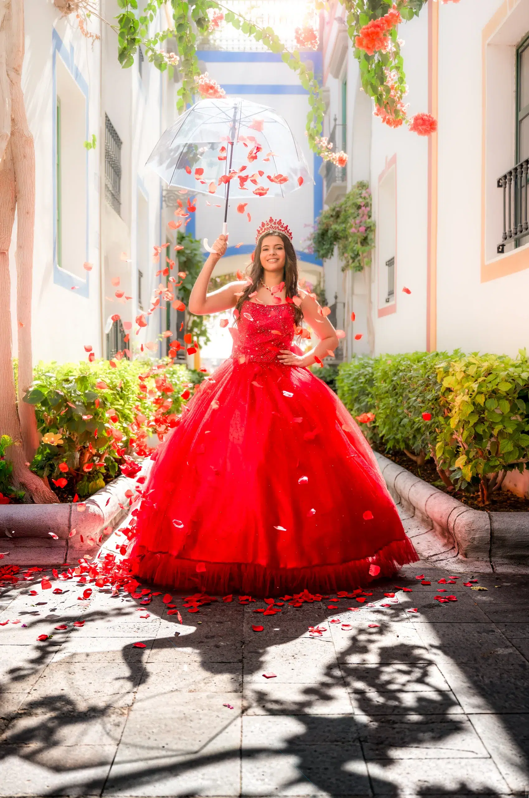 Quinceañera con corona y vestido rojo sosteniendo un paraguas transparente entre pétalos de rosa en Gran Canaria