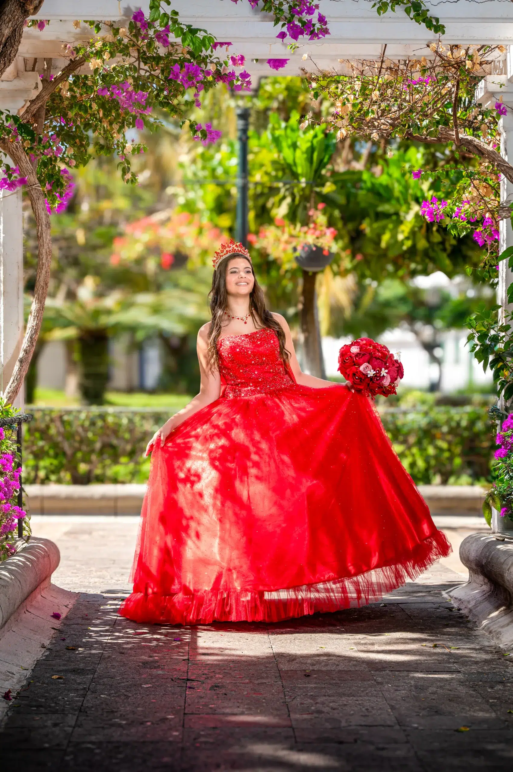 Quinceañera con corona, vestido rojo y ramo de rosas posando bajo una pérgola con buganvillas en Gran Canaria