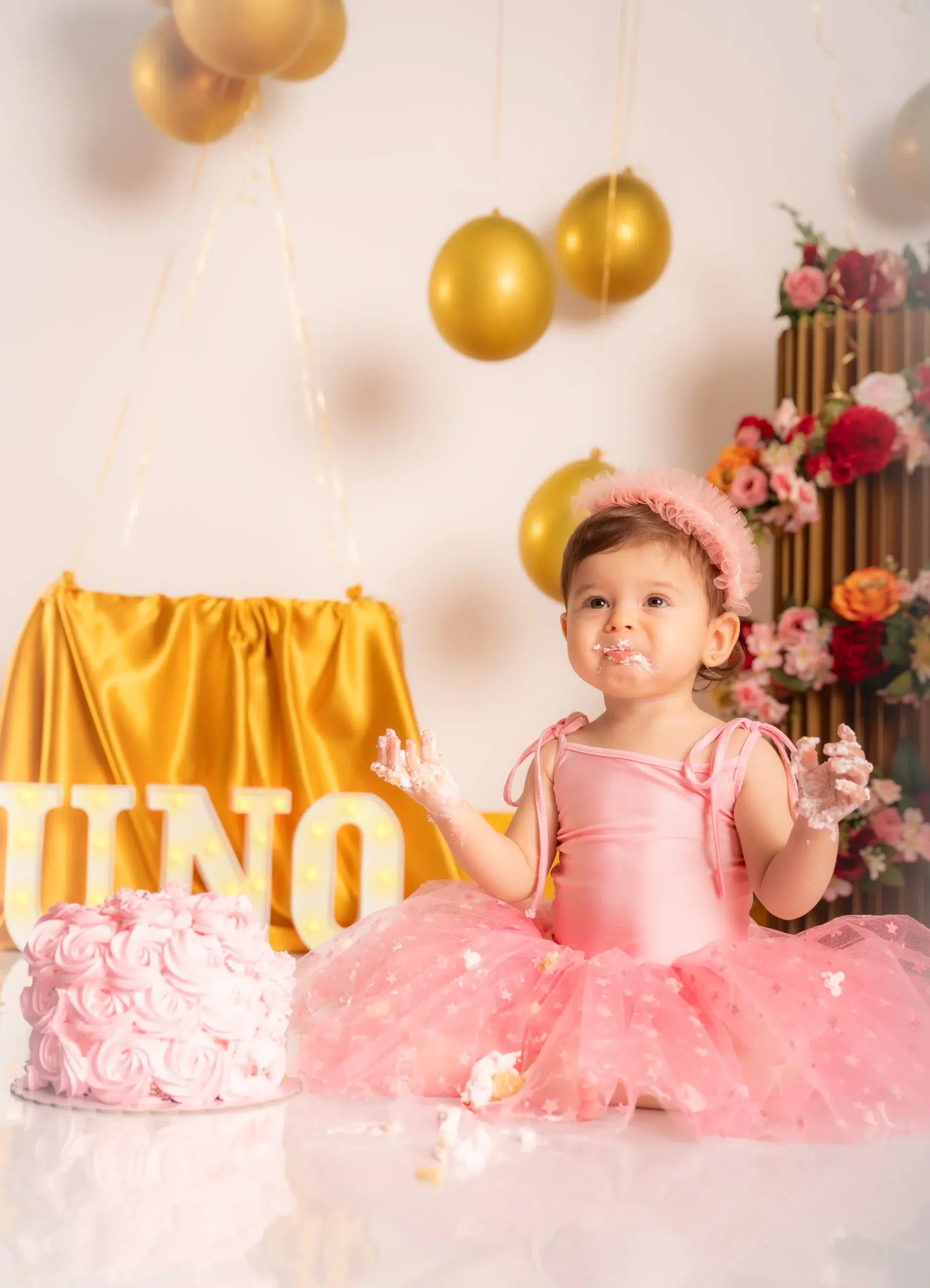 Niña con vestido rosa y las manos cubiertas de crema junto a su tarta de primer cumpleaños en una sesión Smash Cake